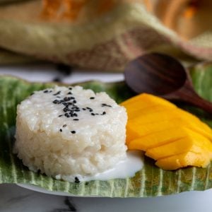 Mango Sticky rice on a banana leaf with a wooden spoon on the side and sprinkled with black sesame seeds.
