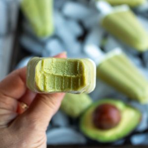 Big bite out of a avocado ice candy. In the background a tray of popsicles and avocado half.