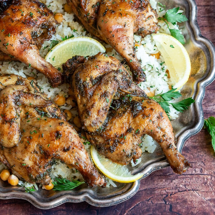 4 little Cornish hen halves on a sliver plate garnished with lemon and fresh parsley.