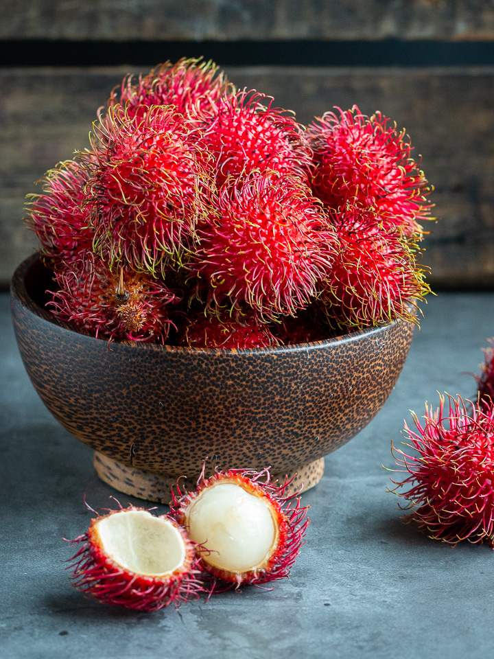 Large wooden bowl filled with fresh rambutan fruits and one is opened to show the snowy white fruit inside.