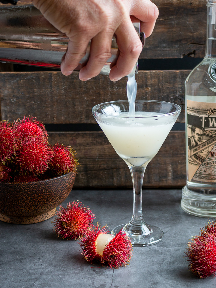 Vodka martini getting poured into a martini glass and rambutans around the table.