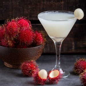 Icy cold Tropical Fruit Rambutan Cocktail in a martini glass with a bowl of fresh rambutans in a wooden bowl.