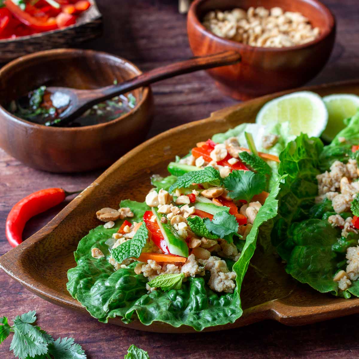 Healthy chicken lettuce wrap topped with fresh vegetables, herbs, roasted peanuts of a wooden plate with prik nam pla sauce next to it.