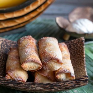 A stack of crispy dessert spring roll in a wooden bowl with a side of whipped coconut cream.