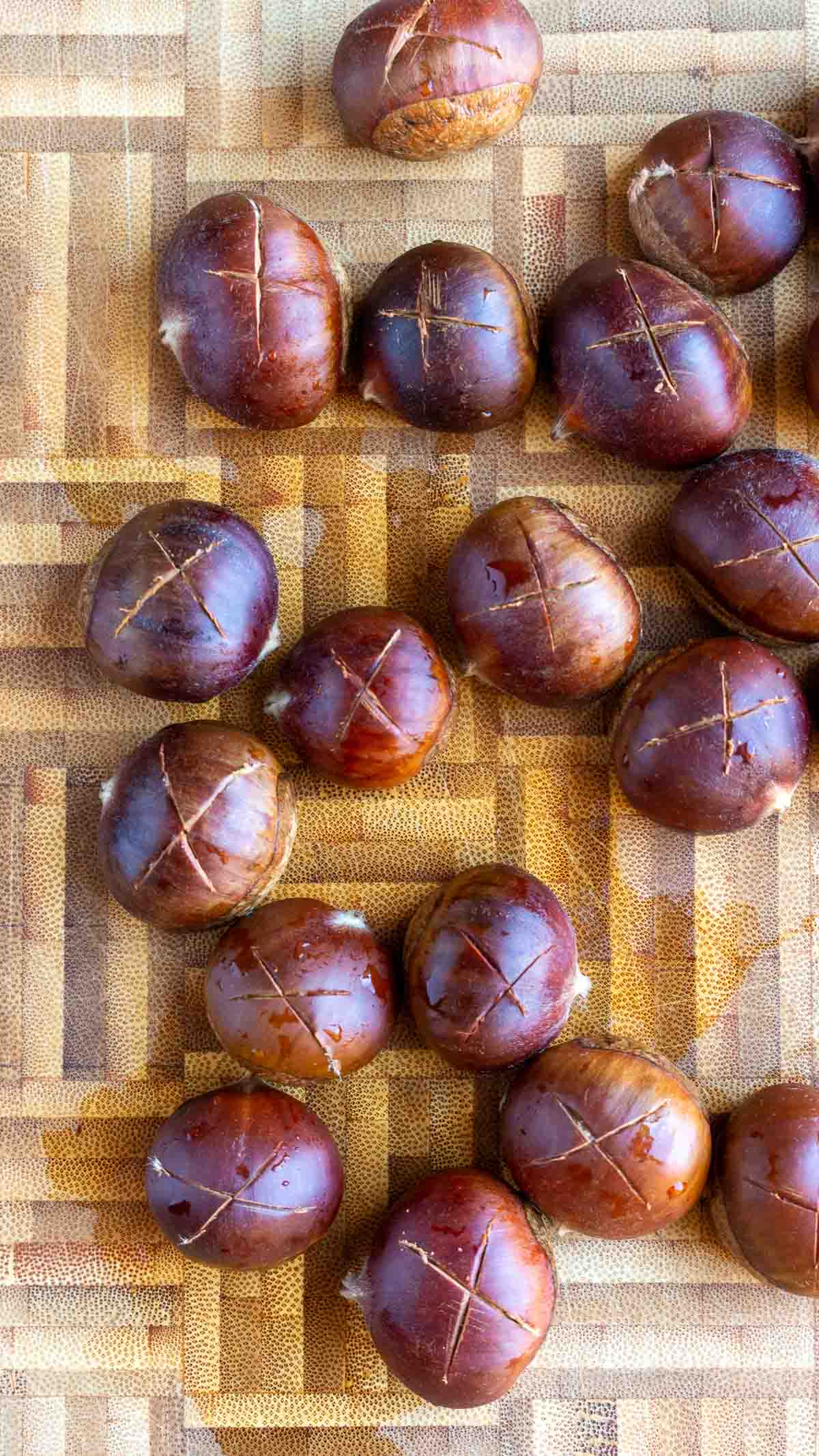 Raw chestnuts with a score of an "x" on the rounded side on a cutting board. 