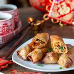 Air fryer spring rolls on a white plate with one cut in half and tea and Chinese New Year decorations in the back.