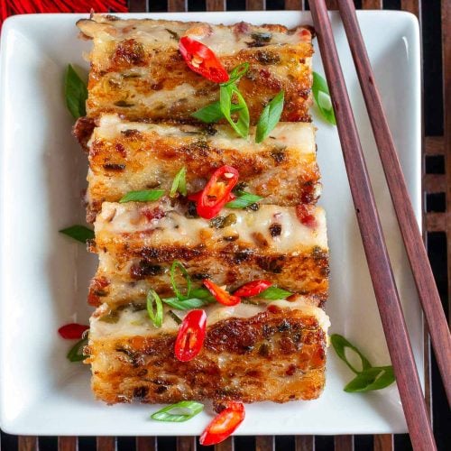 Pan-fried daikon bacon radish cake on a white plate garnished with chilis and green onions with chopsticks.