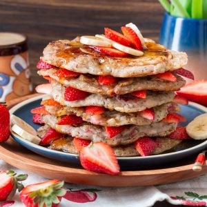 Stacked high strawberry banana pancakes with strawberries between layers on a blue plate with a cup of coffee in the background.