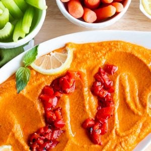 Gluten-free roasted red pepper hummus in a white bowl garnished with roasted red pepper and served with fresh veggies on the side.