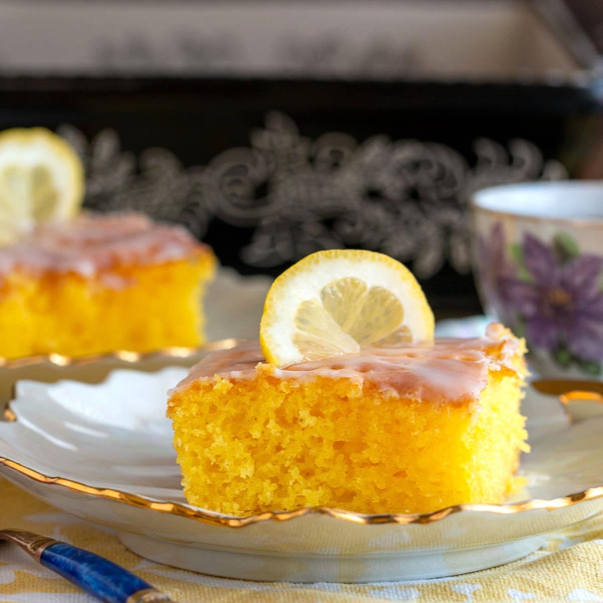 Slice of lemon poke cake with lemon glaze on a white plate served with tea.