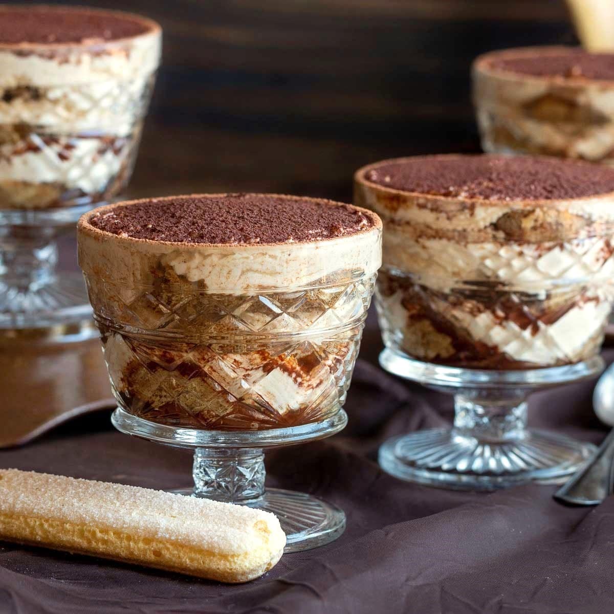 4 mini individual eggless tiramisu cups dusted with cocoa powder with lady finger biscuits on the side.