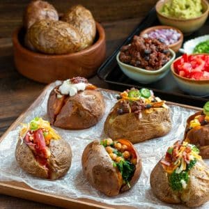 The ultimate baked potato bar is set up with toppings and 6 loaded potatoes on a wooden board.