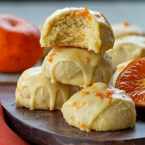 Italian orange drop cookies with a sweet orange icing stacked on a brown plate with a bite out of one. showing the chewy center.