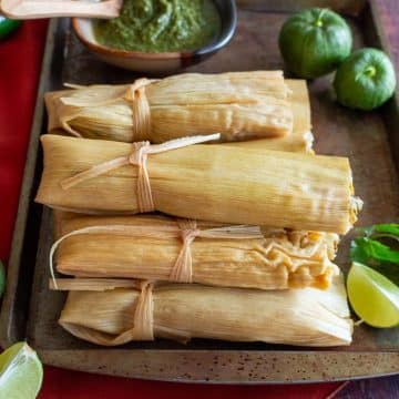 Lots of vegetarian tamales verde stacked up on a brown plate with extra roasted tomatillo salsa on the side.