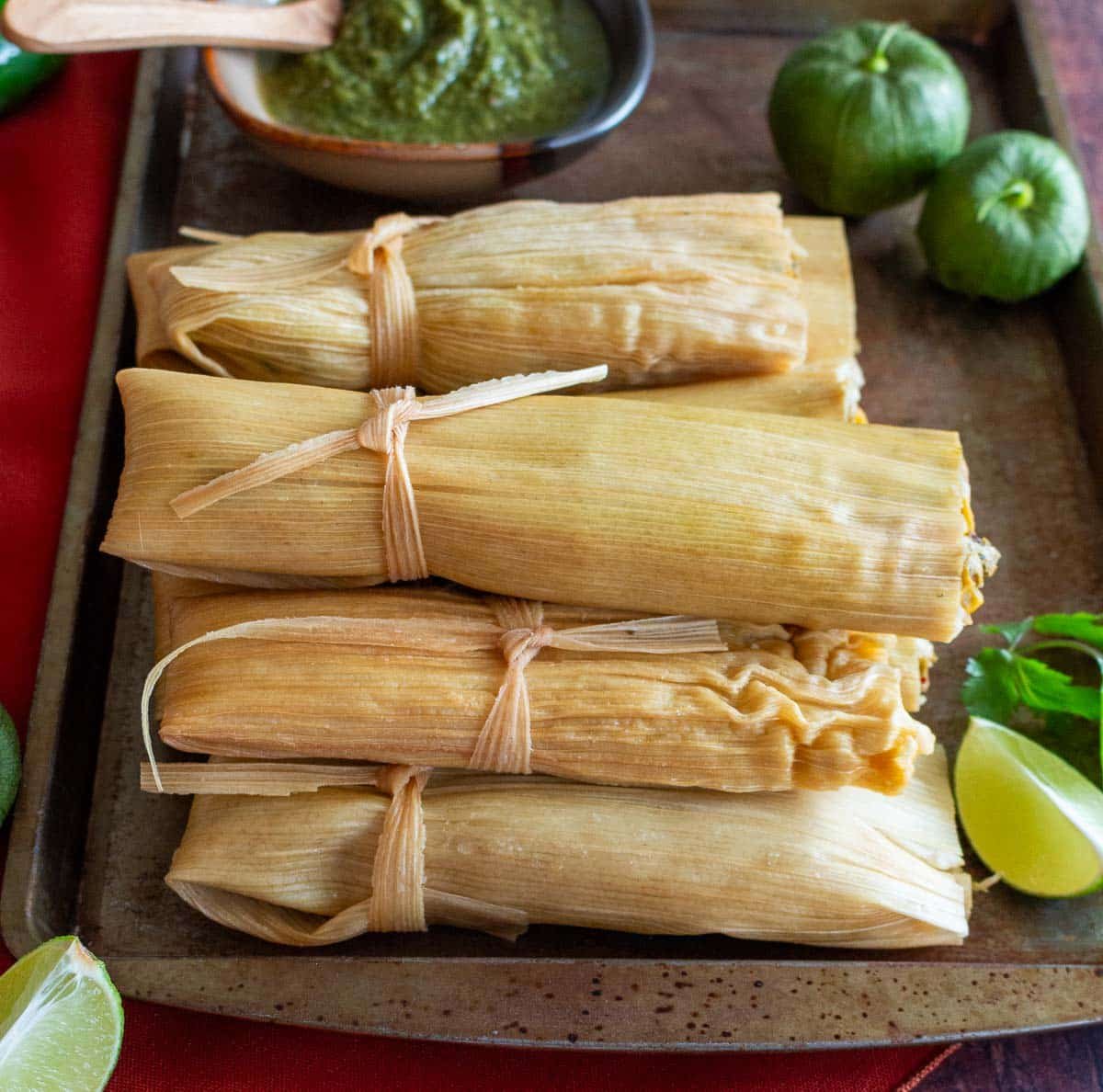 Lots of vegetarian tamales verde stacked up on a brown plate with extra roasted tomatillo salsa on the side.