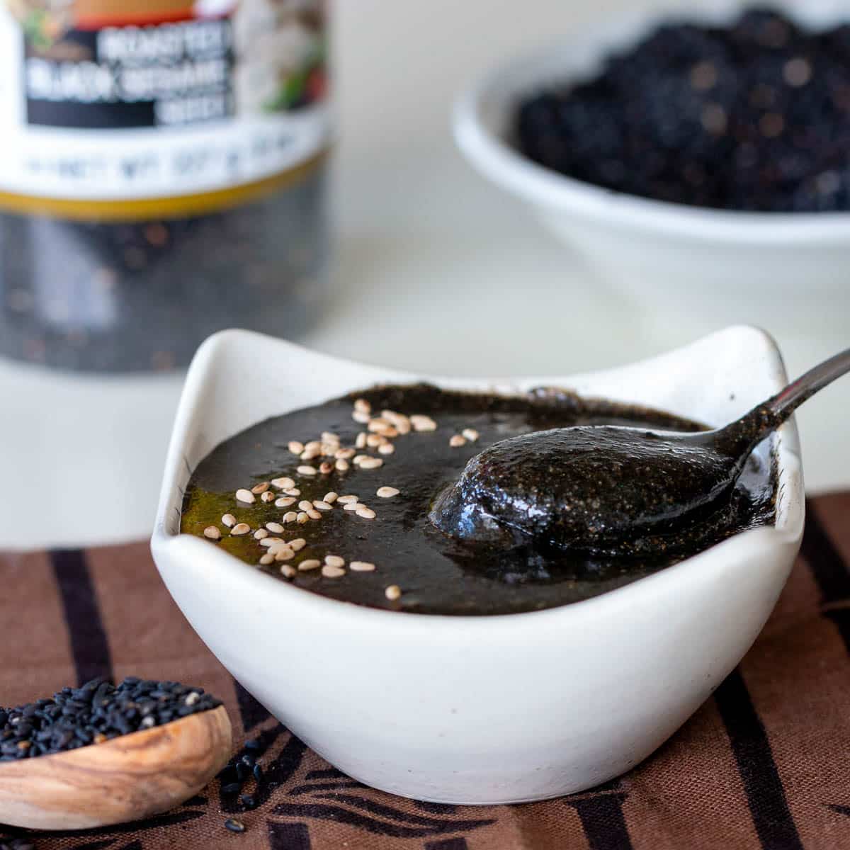 Black seed butter in a white bowl with a spoon and white sesame seeds sprinkled on top.