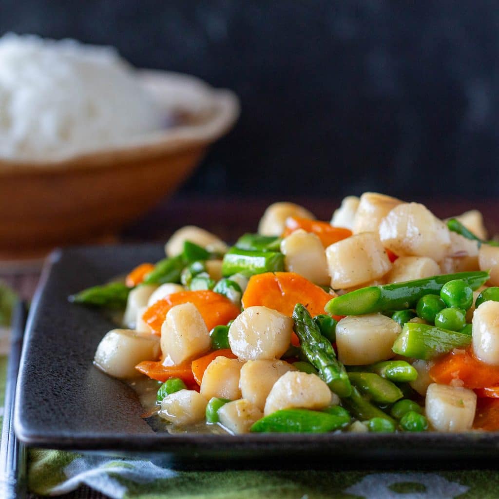Asian bay scallops stir fry with spring vegetables on a gray plate with rice on the side.