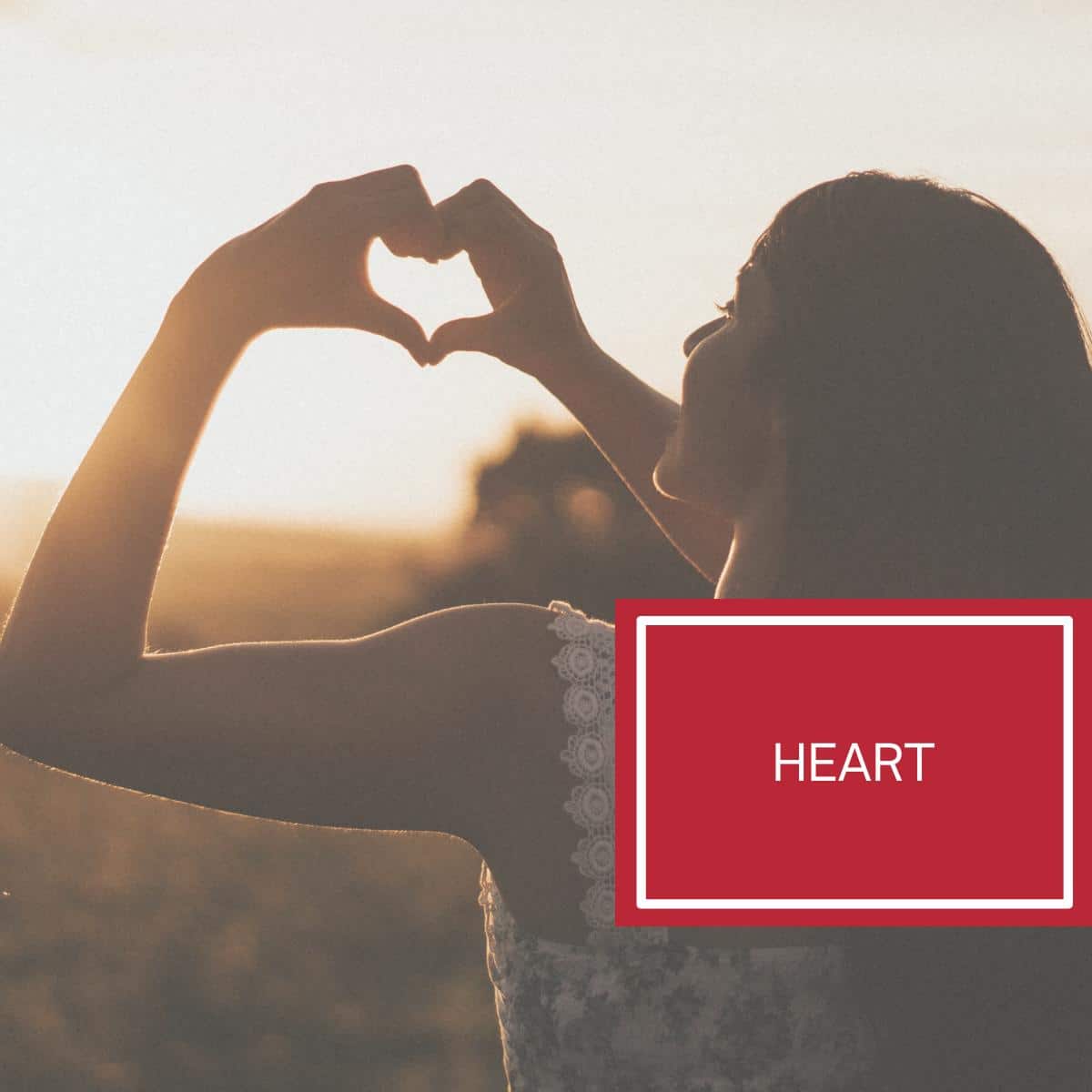 A woman making a heart shape with her hands above the sunset. 