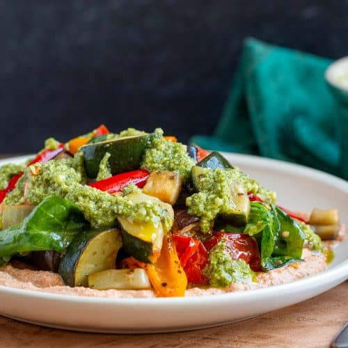 Roasted Italian Vegetables over a creamy whipped tofu ricotta and topped with basil pesto on a white plate.