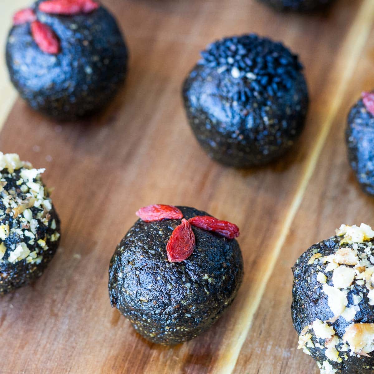 Black sesame bliss balls garnished with walnuts, toasted crushed walnuts and goji berries on a wooden platter.