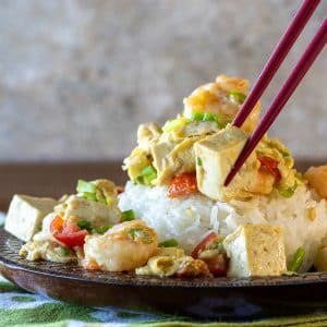 Shrimp and egg stir fry over a bed of steamed white rice grabbing a chunk of tofu with red chopsticks.